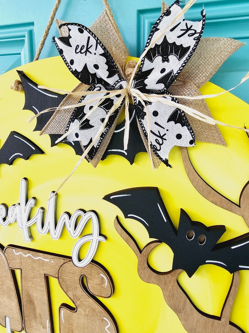Op de afbeelding: Een gele houten deurhanger met de woorden "wreaking havoc" in wit schrift. De deurhanger is versierd met een zwart-witte vleermuis-thema strik, zwarte vleermuizen en bruine takken.