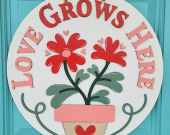 May include: A round, white wooden sign with the words "LOVE GROWS HERE" in red and pink lettering. The sign features a red flower design with green leaves and a pink pot, with a heart detail. A jute rope is attached for hanging.