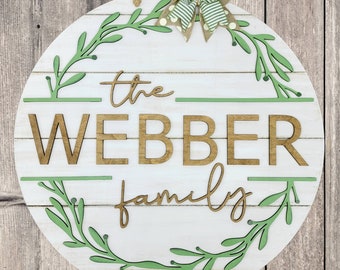 Può includere: Un cartello rotondo in legno con sfondo bianco e un design a corona verde. Il cartello dice "the WEBBER family" in lettere dorate.