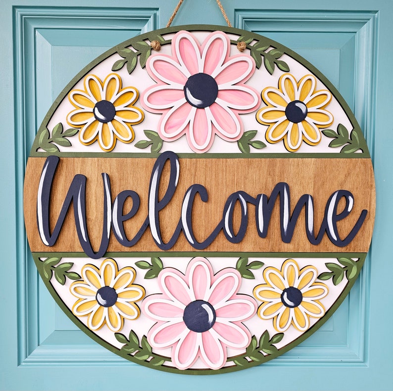 K&ouml;nnte beinhalten: Ein rundes, h&ouml;lzernes Willkommensschild mit wei&szlig;em Hintergrund und gr&uuml;nem Rand. Das Schild zeigt ein Blumenmuster mit rosa, gelben und wei&szlig;en Blumen. Das Wort "Willkommen" ist in schwarzen Buchstaben auf einem braunen Holzpaneel geschrieben.