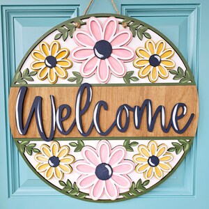 K&ouml;nnte beinhalten: Ein rundes, h&ouml;lzernes Willkommensschild mit floralem Design. Das Schild ist in den Farben Rosa, Gelb und Gr&uuml;n bemalt. Das Wort "Willkommen" ist in schwarzen Buchstaben auf dem Schild geschrieben.