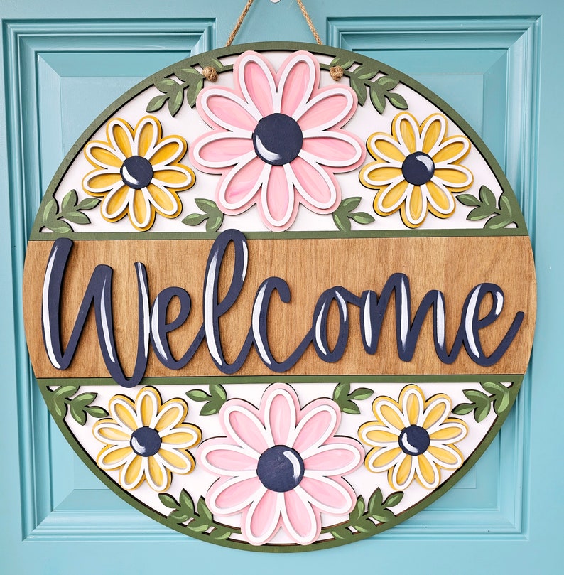 K&ouml;nnte beinhalten: Ein rundes, h&ouml;lzernes Willkommensschild mit floralem Design. Das Schild ist in den Farben Rosa, Gelb und Gr&uuml;n bemalt. Das Wort "Willkommen" ist in schwarzen Buchstaben auf dem Schild geschrieben.