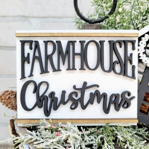 May include: White wooden sign with black lettering that reads "Farmhouse Christmas". The sign is sitting on a wooden surface with a green and white garland.