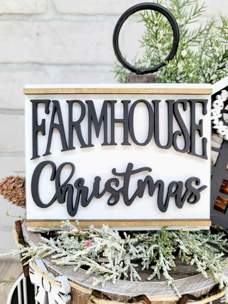May include: White wooden sign with black lettering that reads "Farmhouse Christmas". The sign is sitting on a wooden surface with a green and white garland.