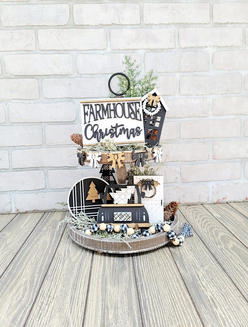May include: A two-tiered wooden tray with a black and white farmhouse Christmas theme. The top tier features a sign that reads "Farmhouse Christmas" and a small wooden house with a wreath. The bottom tier has a black and white patterned circle, a small wooden tree, and a wooden sign with a bow. The tray is decorated with a black and white gingham ribbon and a beaded garland.