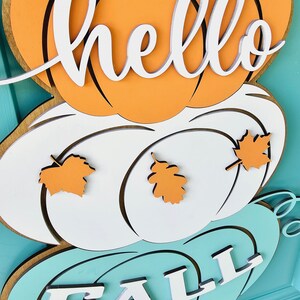 Op de afbeelding: Een houten deurhanger met drie pompoenen. De bovenste pompoen is oranje en heeft het woord "hello" erop geschreven. De middelste pompoen is wit en heeft twee oranje bladeren. De onderste pompoen is turquoise en heeft het woord "fall" erop geschreven.