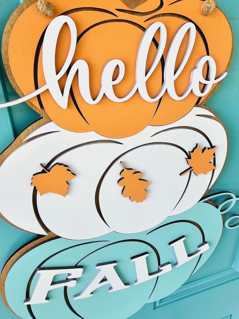 Op de afbeelding: Een houten deurhanger met drie pompoenen. De bovenste pompoen is oranje en heeft het woord "hello" erop geschreven. De middelste pompoen is wit en heeft twee oranje bladeren. De onderste pompoen is turquoise en heeft het woord "fall" erop geschreven.