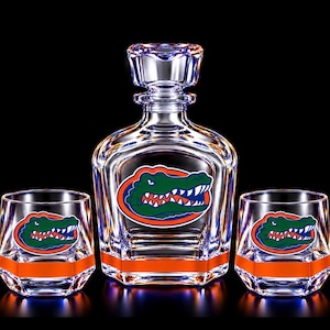 May include: A clear glass decanter with a matching stopper and two matching lowball glasses. Each piece features a green and orange alligator head logo and a horizontal orange band near the base. The glassware has a faceted, geometric design.