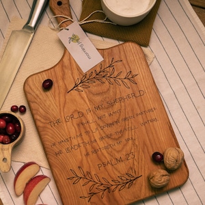 May include: Wooden cutting board with a handle, engraved with Psalm 23. The board is surrounded by ingredients like apples, cranberries, and walnuts. A knife and a small bowl of white powder are also present.