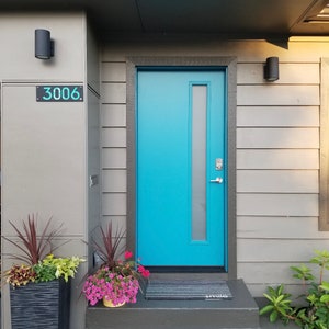 May include: A modern home exterior featuring a vibrant turquoise front door with a vertical glass panel. The door is set in a gray frame, complemented by gray siding and a dark gray roof. A welcome mat reads "smile". Decorative planters with flowers and foliage add color.