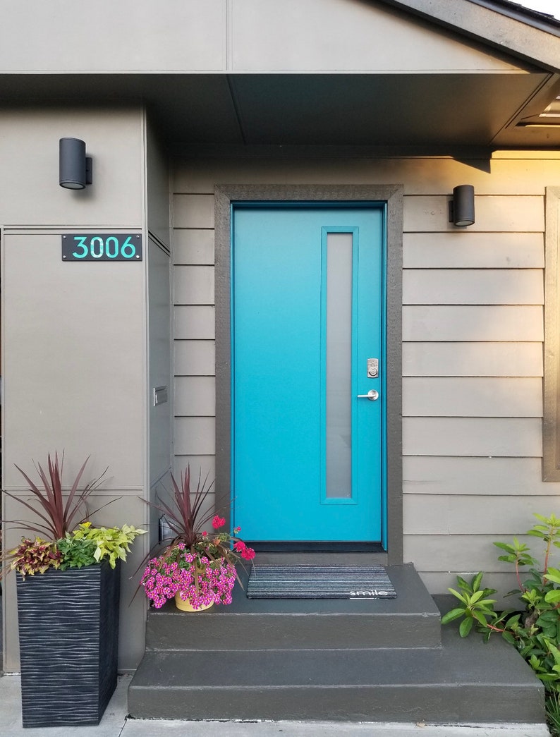 May include: A modern home exterior featuring a vibrant turquoise front door with a vertical glass panel. The door is set in a gray frame, complemented by gray siding and a dark gray roof. A welcome mat reads "smile". Decorative planters with flowers and foliage add color.
