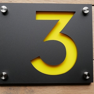 May include: A modern house number sign featuring the number 3. The sign has a black rectangular frame with a yellow backing, and is mounted with four silver screws. The sign is displayed against a wooden background.