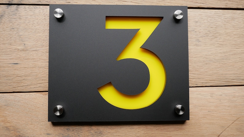 May include: A modern house number sign featuring the number 3. The sign has a black rectangular frame with a yellow backing, and is mounted with four silver screws. The sign is displayed against a wooden background.