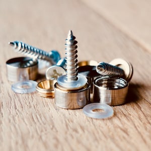 May include: Close-up of various metal hardware components, including screws, cylindrical caps, and clear plastic washers. The hardware is silver and gold-toned, arranged on a light brown wooden surface. The components are for construction or assembly.