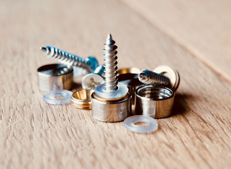 May include: Close-up of various metal hardware components, including screws, cylindrical caps, and clear plastic washers. The hardware is silver and gold-toned, arranged on a light brown wooden surface. The components are for construction or assembly.