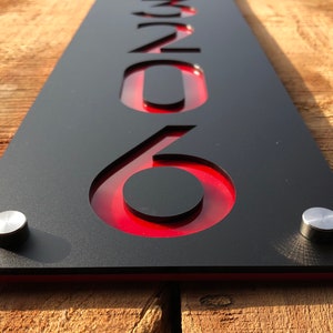 May include: Black house number sign with red acrylic inlay, featuring the number 206. The sign is mounted on a wooden surface with silver screws.