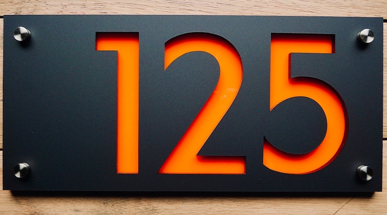 May include: A modern house number sign featuring the number "125" cut out of a black rectangular plate. The numbers are filled with an orange color, and the sign is mounted with silver screws on a wooden surface.