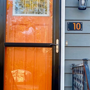 May include: An orange front door with a black frame and a small window with a grid pattern. A black rectangular house number plaque displays the number "10" in orange. The door has a silver handle.