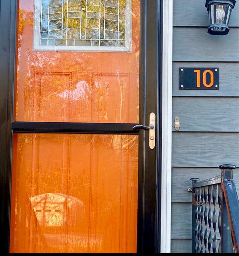 May include: An orange front door with a black frame and a small window with a grid pattern. A black rectangular house number plaque displays the number "10" in orange. The door has a silver handle.