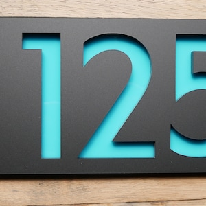 May include: A modern black house number sign with the numbers "125" cut out, revealing a turquoise backing. The sign is mounted with silver hardware on a wooden surface.