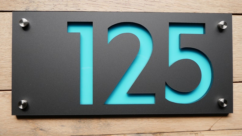 May include: A modern black house number sign with the numbers "125" cut out, revealing a turquoise backing. The sign is mounted with silver hardware on a wooden surface.