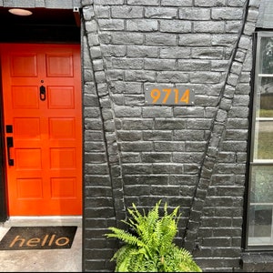 May include: An orange front door with a black door handle and a welcome mat. The door is next to a black brick wall with the number 9714 in orange. A green fern plant is at the base of the wall.