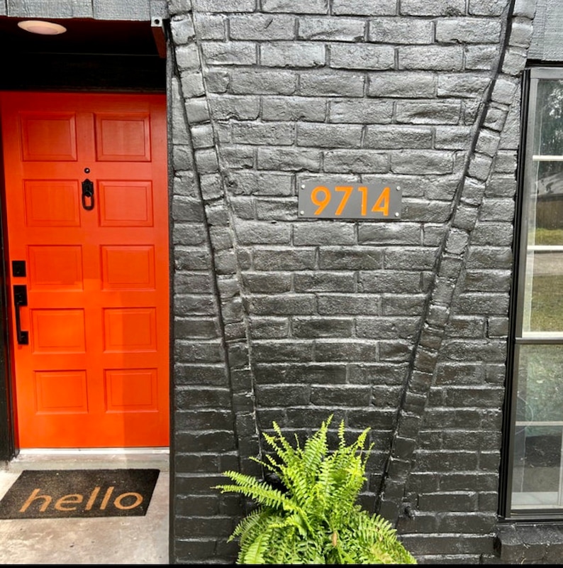 May include: An orange front door with a black door handle and a welcome mat. The door is next to a black brick wall with the number 9714 in orange. A green fern plant is at the base of the wall.