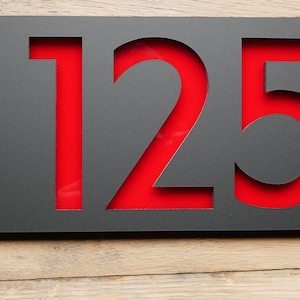 May include: A modern house number sign featuring the number "125" cut out of a dark gray metal plate. The numbers are filled with a vibrant red backing. The sign is mounted with four silver screws on a wooden surface.