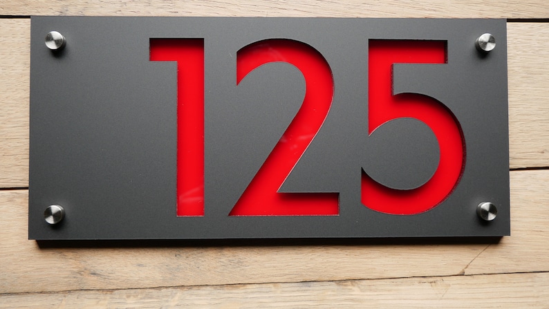 May include: A modern house number sign featuring the number "125" cut out of a dark gray metal plate. The numbers are filled with a vibrant red backing. The sign is mounted with four silver screws on a wooden surface.