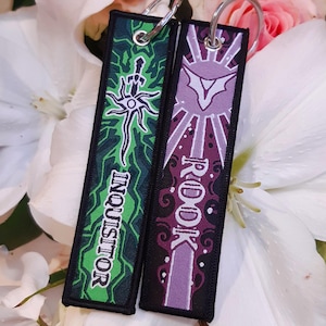 Peut inclure: Deux porte-clés rectangulaires avec des anneaux argentés. L'un est vert avec le mot "INQUISITOR" et un motif blanc et noir. L'autre est violet avec le mot "ROOK" et un motif blanc et violet. Les deux sont sur un fond floral.