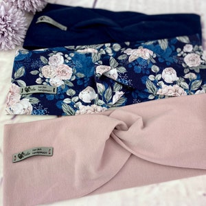 May include: Three fabric headbands, one navy blue, one with a floral pattern in shades of blue and white, and one light pink. Each headband has a small tag that reads "herzlich handgemacht".