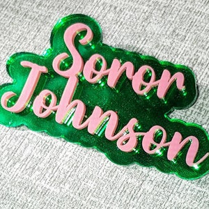 May include: A layered acrylic name tag in green and pink. The name tag features the name "Soror Johnson" in a cursive font, layered over a green base. The name tag has a scalloped edge.