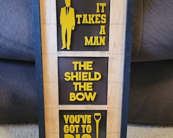 May include: A framed wooden sign with three panels. The top panel has a silhouette of a person in a suit and the text "It takes a man". The middle panel has the text "The shield the bow". The bottom panel has a shovel and the text "You've got to dig".