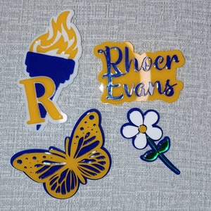 May include: A collection of yellow and blue decorative stickers. The stickers include a torch with flames, the letter "R", a butterfly, a flower, and the name "Rhoer Evans".