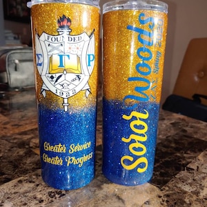 May include: Two gold and blue glitter tumblers. One tumbler features a sorority crest with the letters "ΣΓΡ" and the text "Founded 1922". The other tumbler has the text "Soror Woods 2023" and "Greater Service Greater Progress".