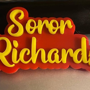 May include: A red and yellow 3D name plaque with the name "Sonor Richards" in yellow lettering.