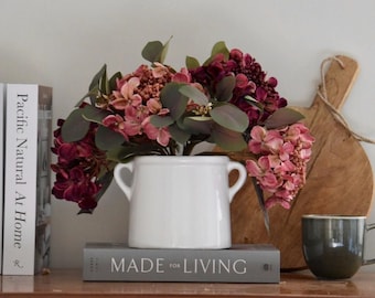 May include: A white ceramic vase with two handles filled with a bouquet of pink and burgundy flowers. The vase is sitting on top of a book titled "Made for Living".