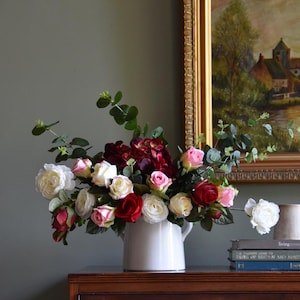 May include: A white ceramic pitcher overflowing with a vibrant arrangement of roses in shades of red, pink, and white, accented with green foliage. The floral display sits atop a dark wooden dresser, with a framed painting in the background.