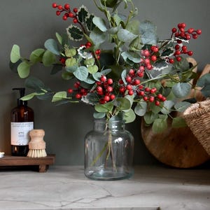 Könnte beinhalten: Eine klare Glasvase enthält ein Arrangement aus künstlichem Grün und roten Beeren. Das Arrangement umfasst Eukalyptusblätter und Stechpalmen, wodurch eine festliche Atmosphäre entsteht. Die Vase steht auf einer Arbeitsplatte.