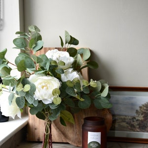 Puede incluir: Un ramo de peon&iacute;as blancas y hojas de eucalipto atadas con una cuerda, sobre una tabla de cortar de madera. Un tarro de vela de vidrio marr&oacute;n con una etiqueta blanca que dice "FRAGRANCED CANDLE -  PINK CARROT, GREEN TEA & AMBER" est&aacute; sobre una encimera de m&aacute;rmol.