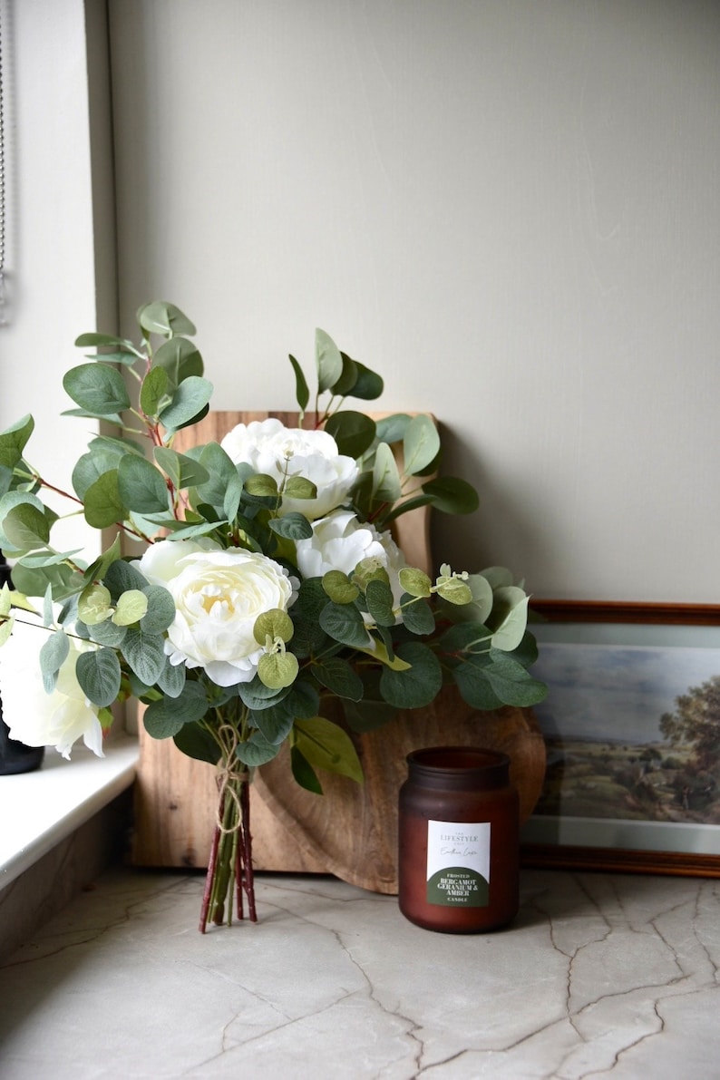 Puede incluir: Un ramo de peon&iacute;as blancas y hojas de eucalipto atadas con una cuerda, sobre una tabla de cortar de madera. Un tarro de vela de vidrio marr&oacute;n con una etiqueta blanca que dice "FRAGRANCED CANDLE -  PINK CARROT, GREEN TEA & AMBER" est&aacute; sobre una encimera de m&aacute;rmol.