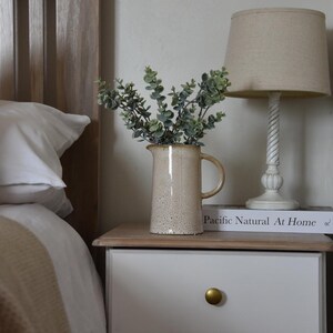 May include: A white bedside table with two drawers and gold knobs. A beige ceramic pitcher with a speckled finish holds a bouquet of green eucalyptus branches. A book titled "Pacific Natural At Home" sits on top of the table, and a beige lamp with a linen shade sits behind the pitcher.