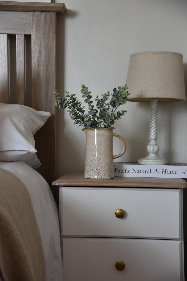 May include: A white bedside table with two drawers and gold knobs. A beige ceramic pitcher with a speckled finish holds a bouquet of green eucalyptus branches. A book titled "Pacific Natural At Home" sits on top of the table, and a beige lamp with a linen shade sits behind the pitcher.