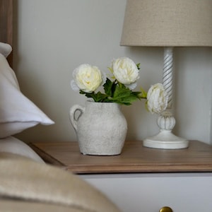 May include: A small, textured, off-white vase with a handle contains white flowers and green leaves. The vase is on a wooden bedside table next to a white lamp with a beige lampshade. The scene suggests a peaceful atmosphere.
