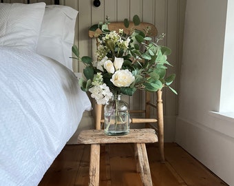 May include: A white bouquet of artificial flowers in a clear glass vase sits on a wooden stool in a bedroom. The flowers are arranged in a vase on a wooden stool in front of a white bed with white bedding.