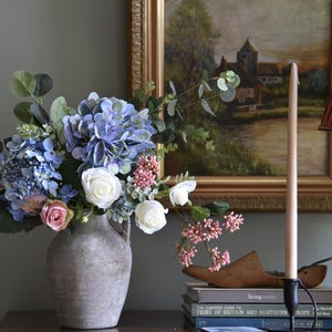 Pu&ograve; includere: Una natura morta con un bouquet di ortensie blu, rose bianche e fiori rosa in un vaso grigio. Un dipinto di paesaggio incorniciato e una candela alta in un supporto nero sono su una superficie di legno.