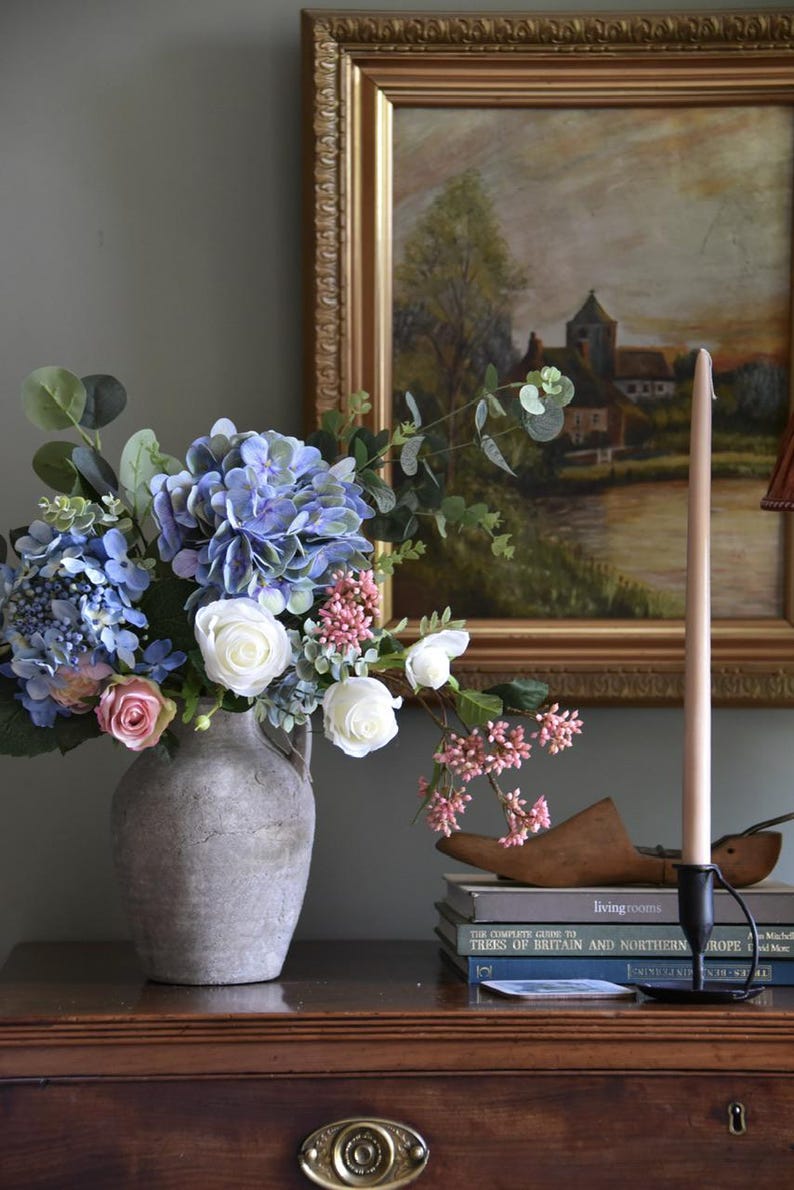 Pu&ograve; includere: Una natura morta con un bouquet di ortensie blu, rose bianche e fiori rosa in un vaso grigio. Un dipinto di paesaggio incorniciato e una candela alta in un supporto nero sono su una superficie di legno.