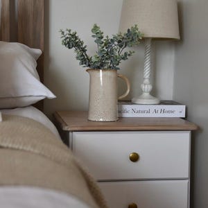 May include: A white nightstand with three drawers and brass knobs. The nightstand has a wooden top with a book and a ceramic pitcher with green leaves on top. A white lamp with a beige lampshade is on the nightstand.