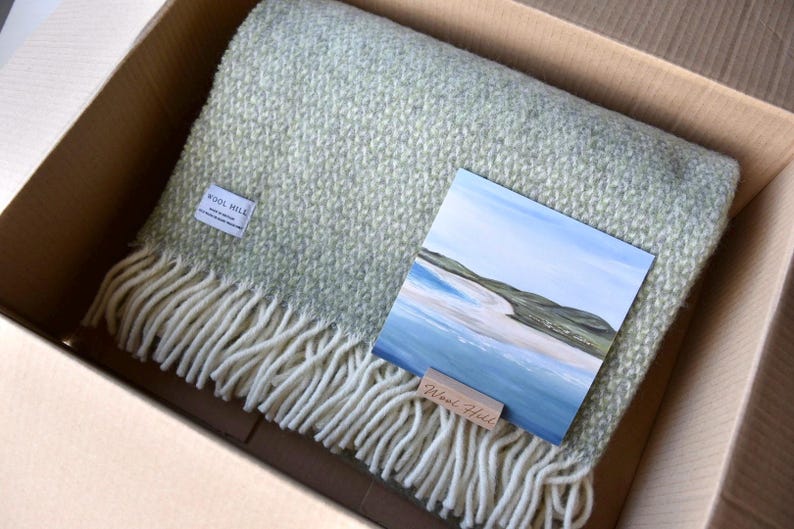 K&ouml;nnte beinhalten: Eine gr&uuml;ne und wei&szlig;e Wolldecke mit Fransenrand ist in einem Karton gefaltet. Auf der Decke liegt eine kleine Karte mit einem Gem&auml;lde einer Strandszene. Die Karte tr&auml;gt den Text "Wool Hill".