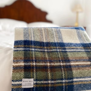 Puede incluir: Una manta de lana a cuadros blancos, azules, verdes y marrones con flecos. La manta tiene una etiqueta que dice "Wool Hill Made in Britain 100% Wool Hand Wash Only".